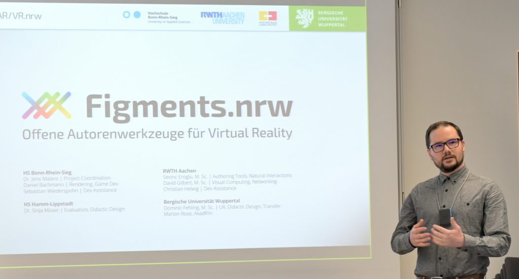 This image shows Dominic Fehling standing in front of a projection screen giving the introduction to the workshop demonstration of Figments.nrw. On the screen is a slide of the presentation visible that is showing the logo of Figments.nrw as well as the names of all the developers and universities involved in the project.